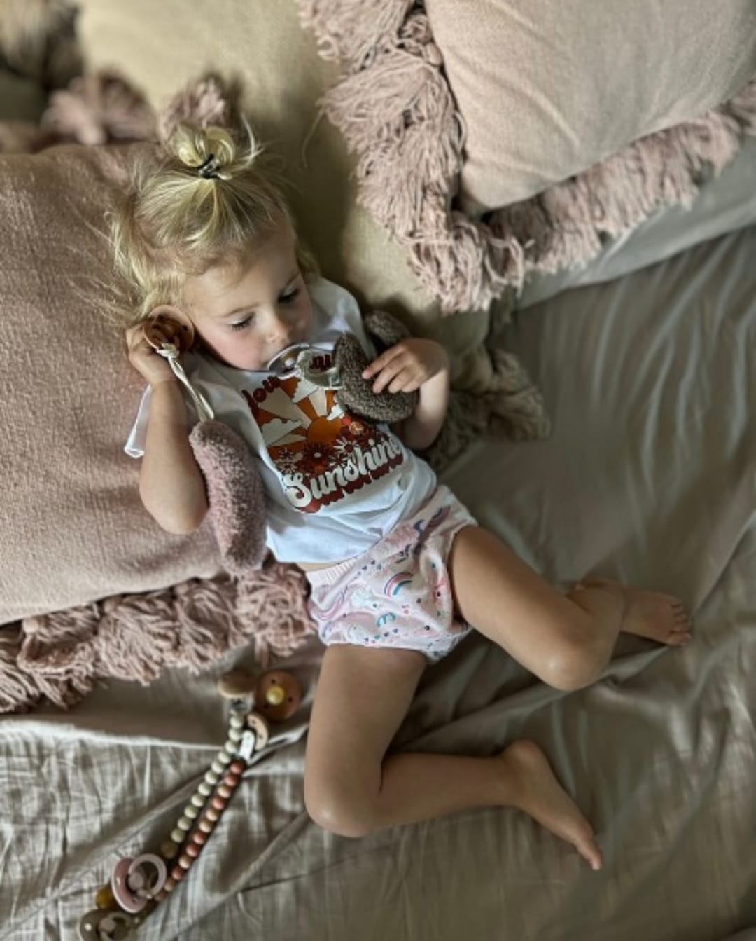 Petite fille allongée sur un lit portant une culotte d’apprentissage lavable à motifs arc-en-ciel, jouant avec une peluche